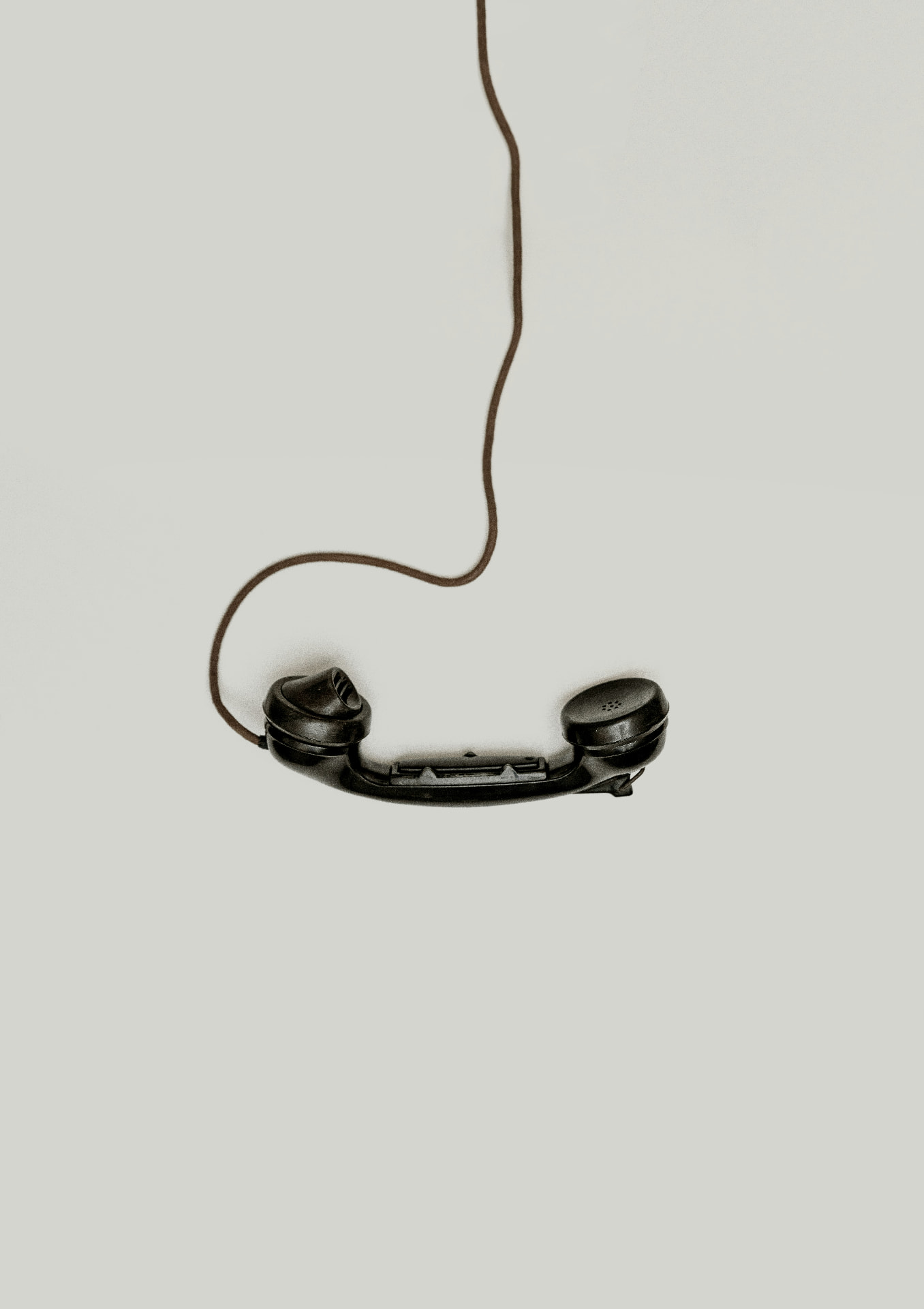 A black telephone receiver hanging in mid-air from a coiled cord, against a pale grey background