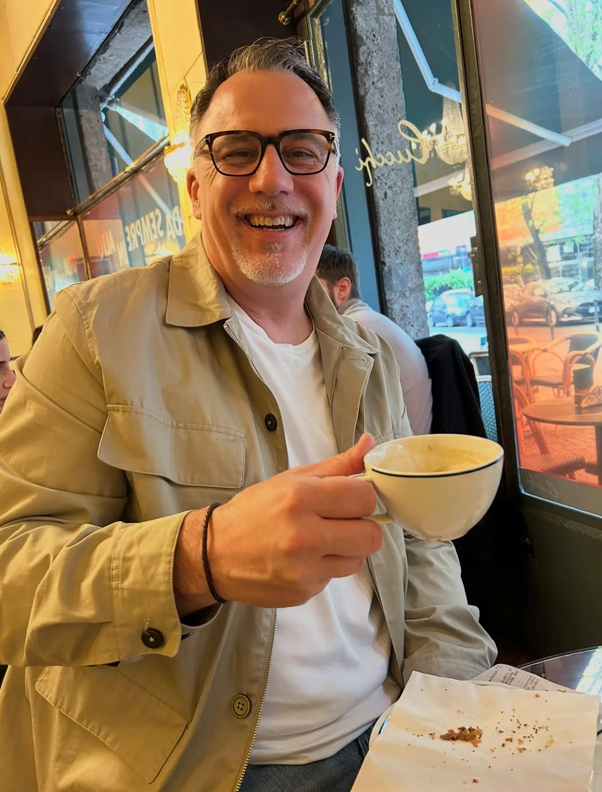 Glenn Issler seated at a café table beside a window, smiling, with a cup of coffee in his hand